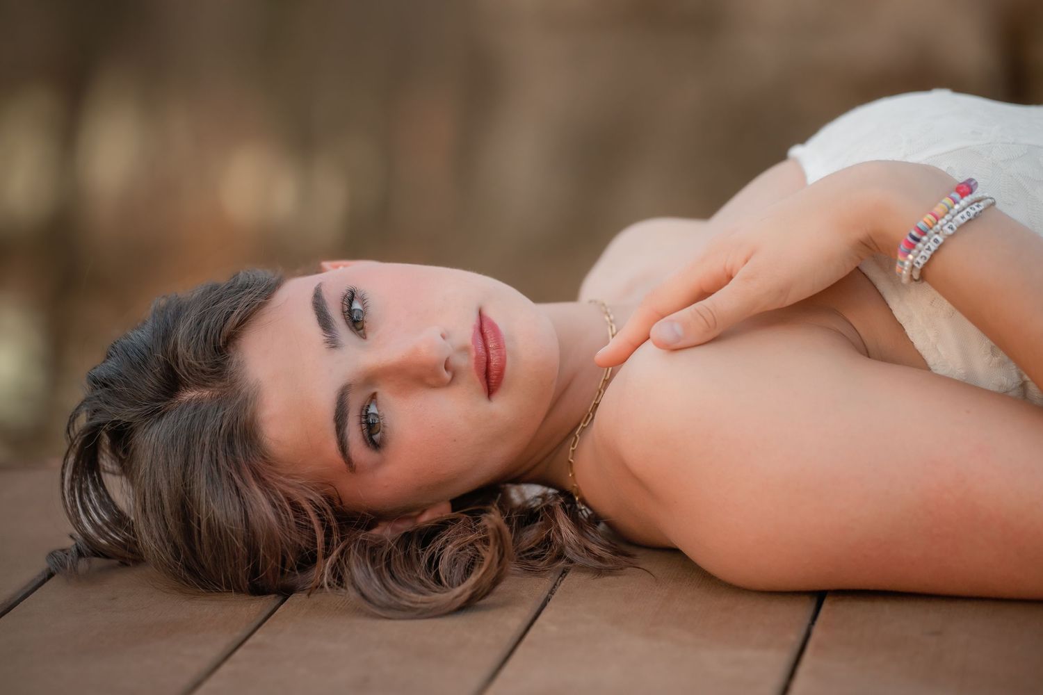 Young woman with curly hair lying on wooden floor wearing white dress and bracelet, gazing at camera.
