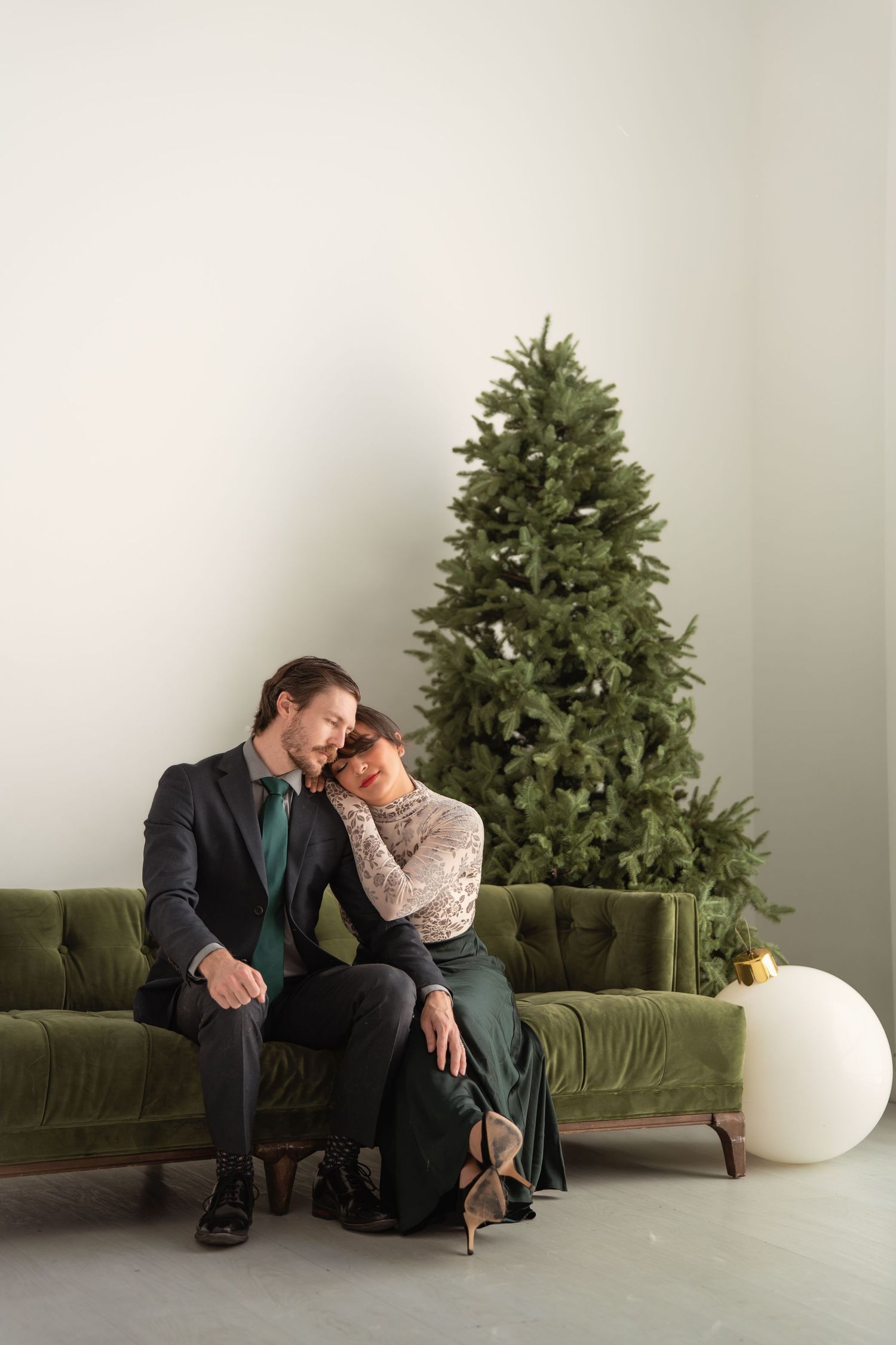 Elegant couple in formal wear relaxing on a green velvet sofa beside an undecorated Christmas tree in a minimalist room.