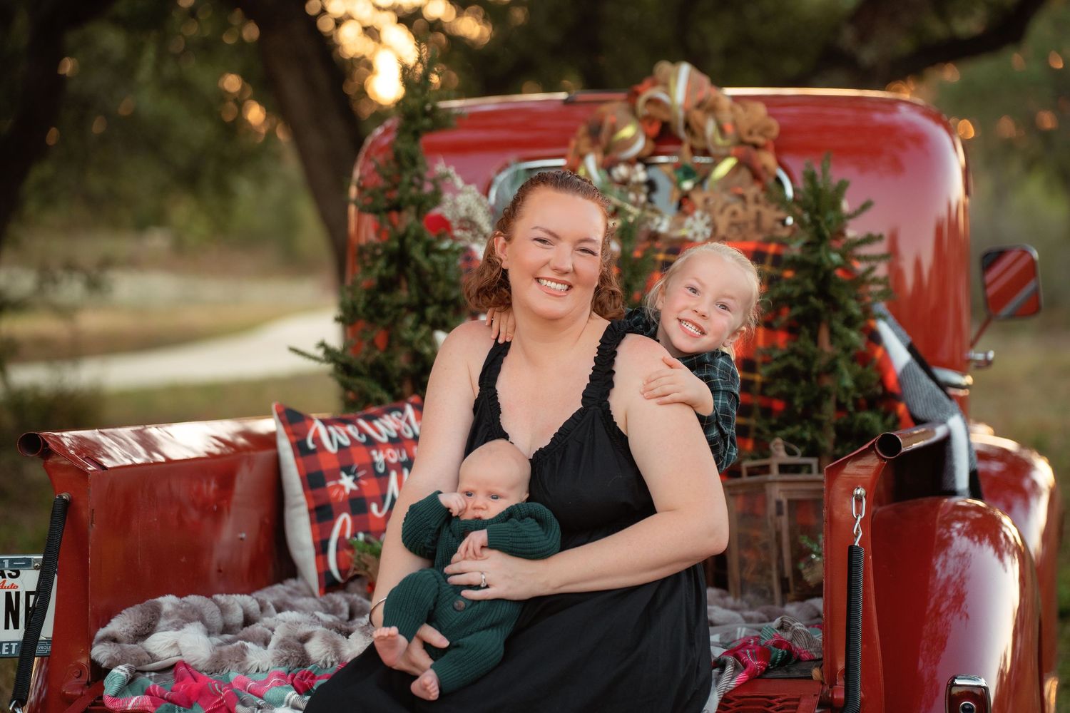 Woman holding newborn baby with toddler beside her, posing in front of a decorated red truck for a Christmas photo.