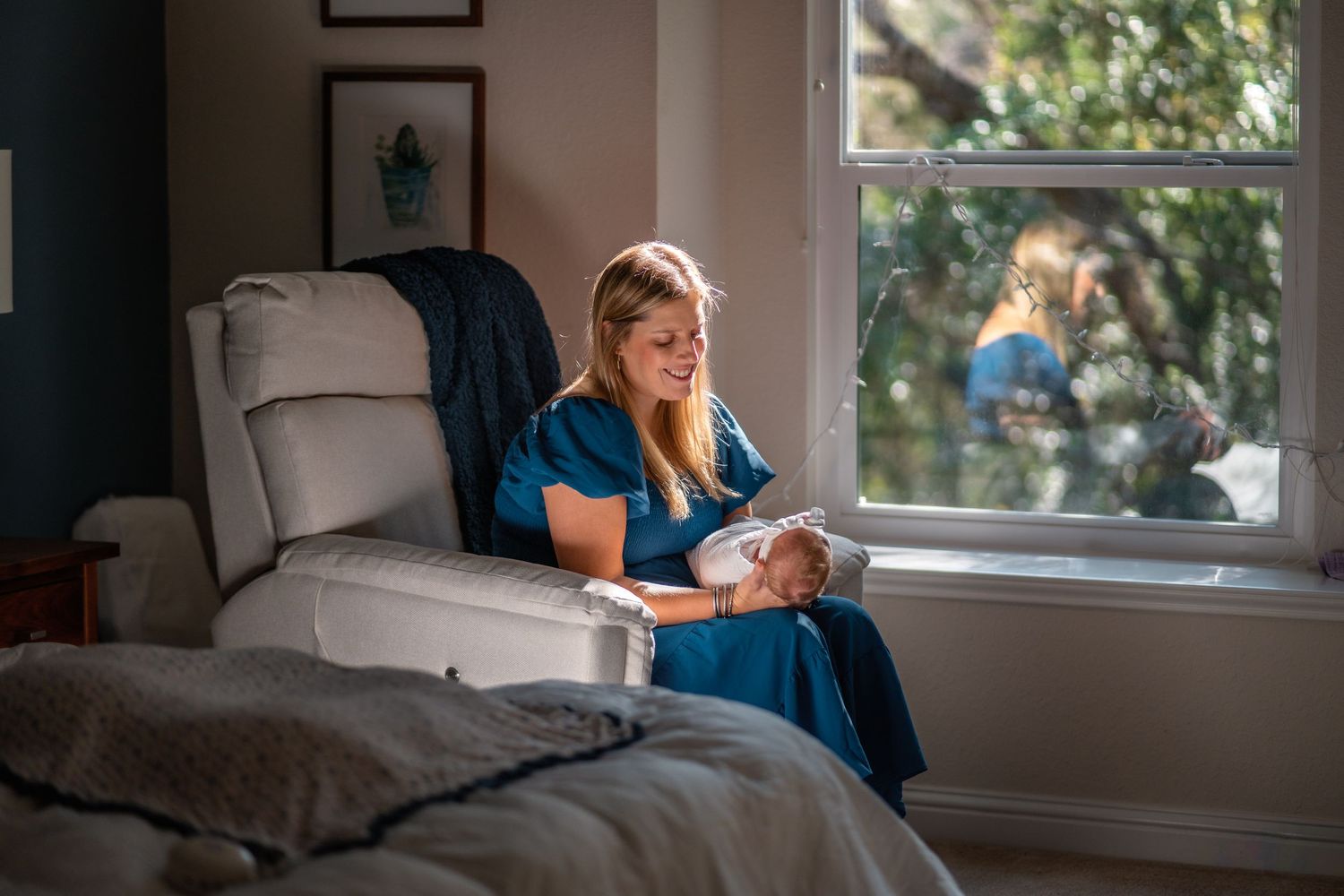 A woman in a blue dress sits in a white recliner by a window, tenderly cradling a newborn baby in soft natural light.