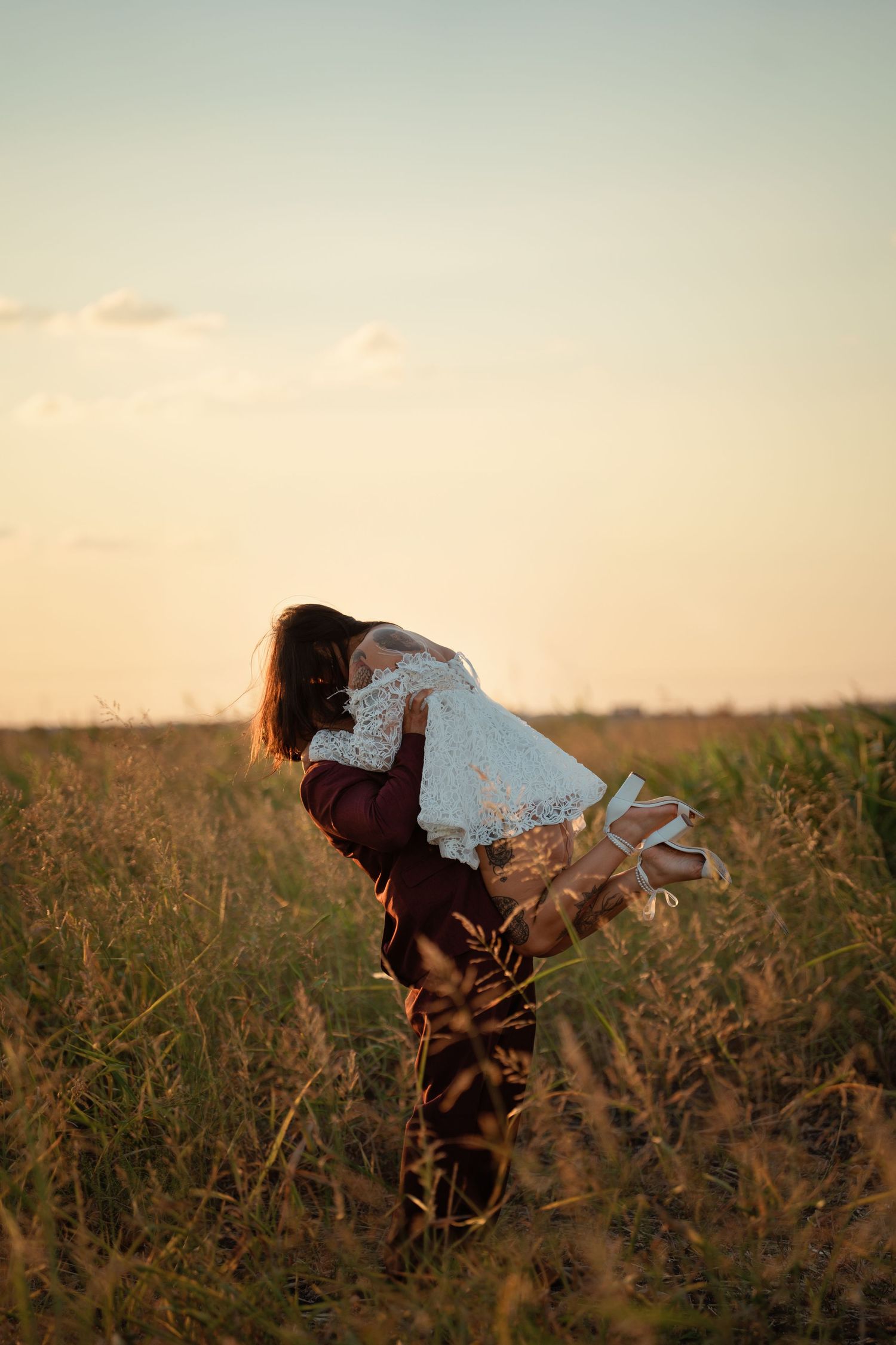 A woman in a burgundy dress holds a white lace garment in a golden field at sunset.