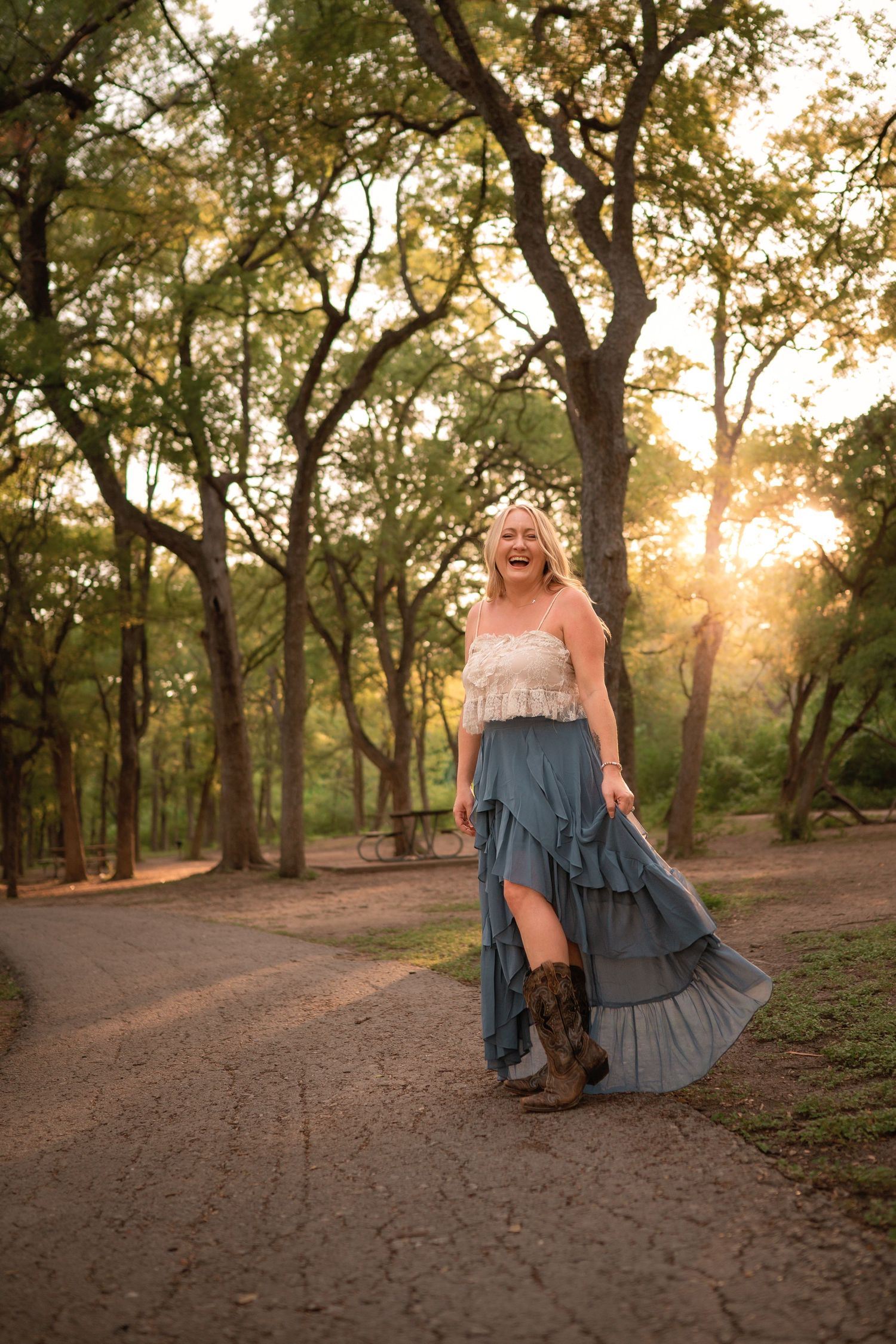 Woman in blue ruffled skirt and cowboy boots laughing on a sunlit forest path at golden hour.