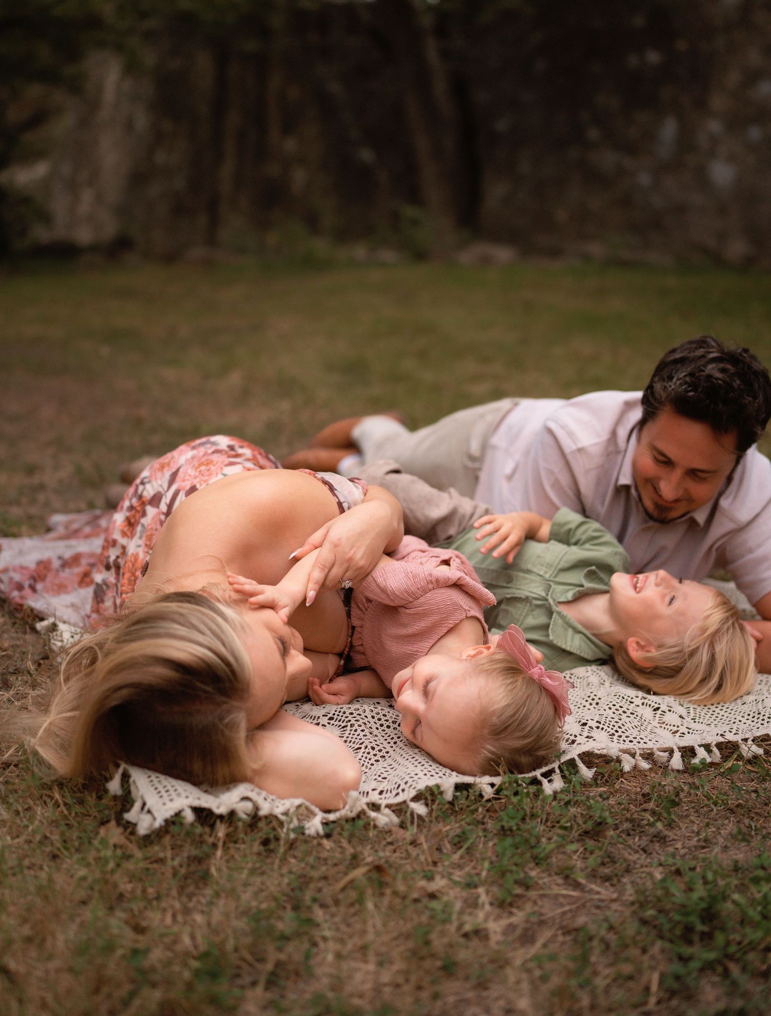 Family of four lying on a crochet blanket outdoors, parents cuddling two young girls on grass near trees.