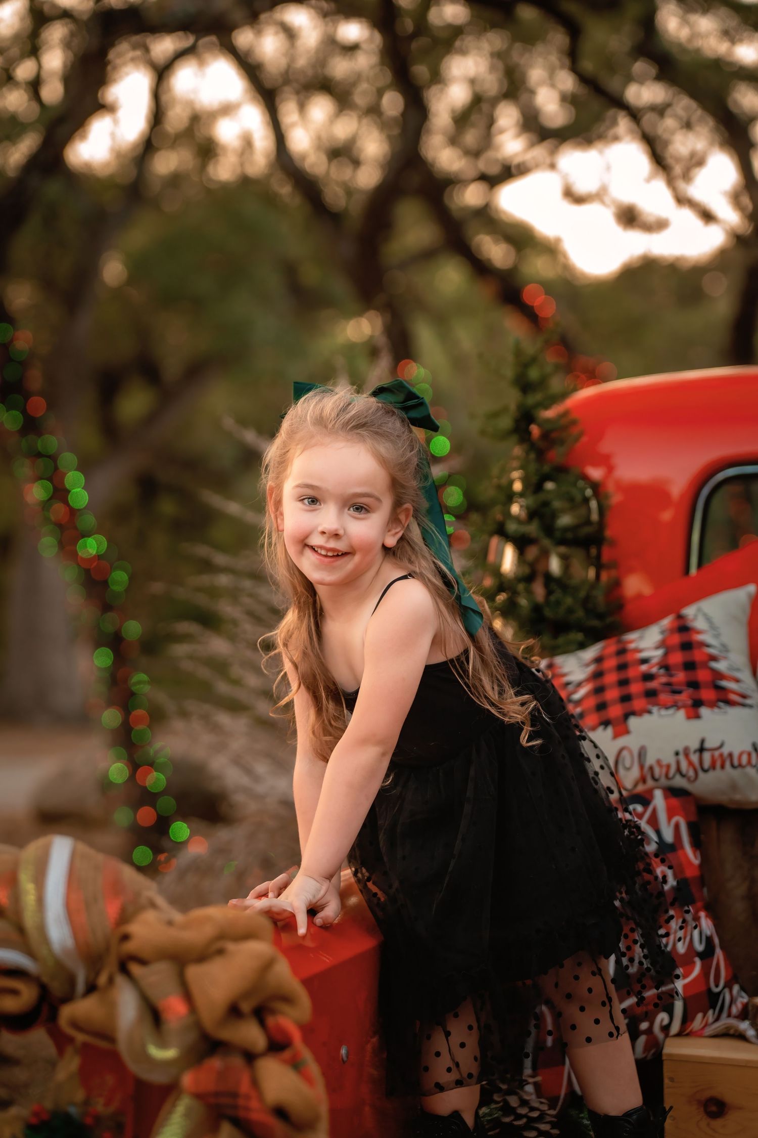Young girl in black tutu dress poses by red Christmas truck with festive lights and holiday decorations outdoors.