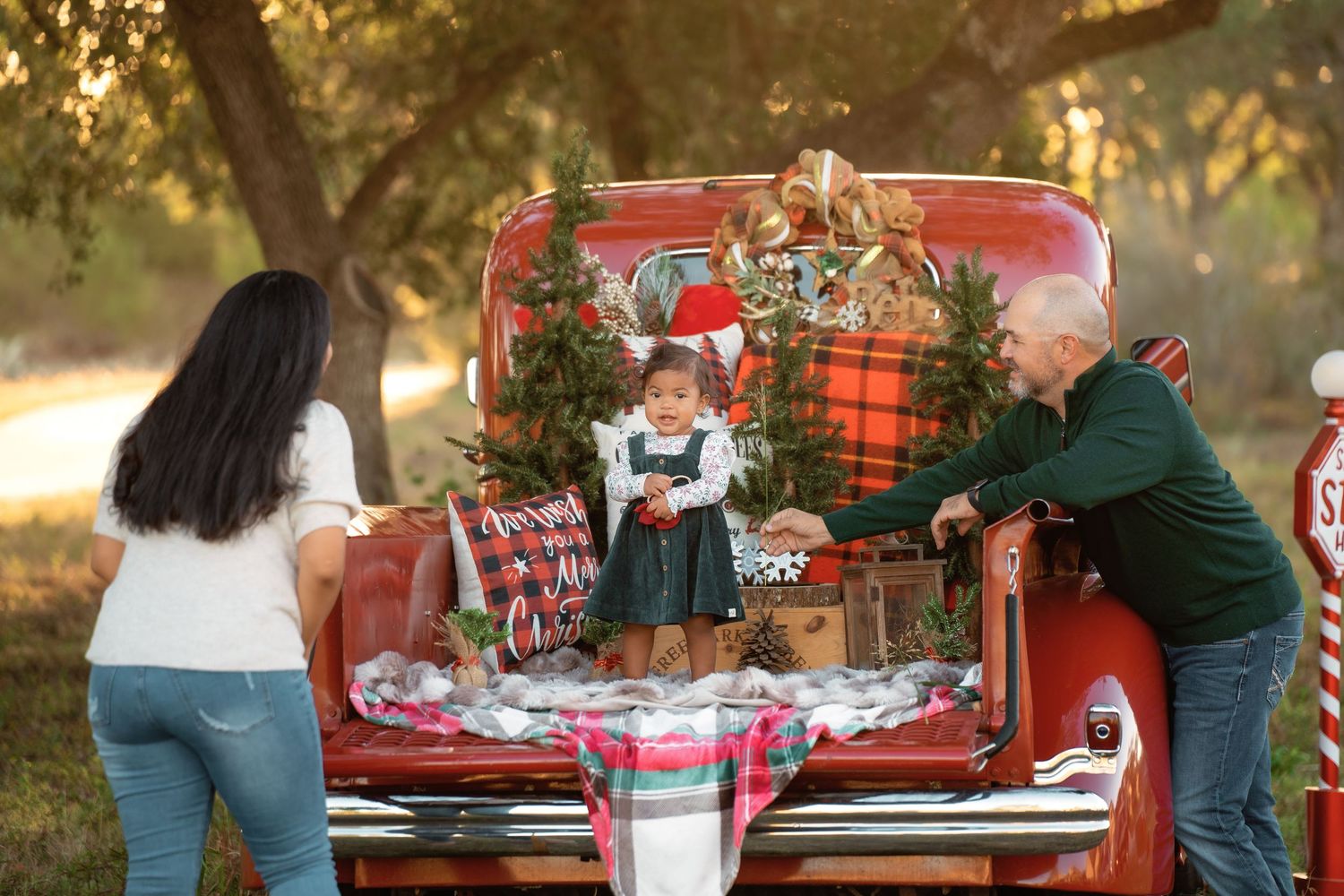 Family Christmas photo shoot with toddler in red truck bed decorated with wreaths and plaid holiday decor outdoors.