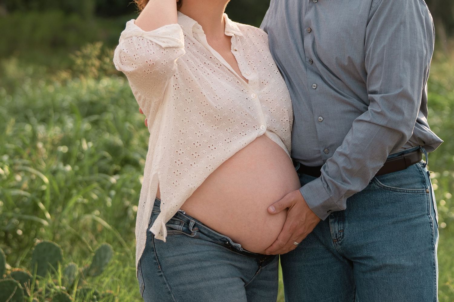 Couple outdoors showing pregnant belly, man tenderly holding woman's baby bump in a green field maternity photo.