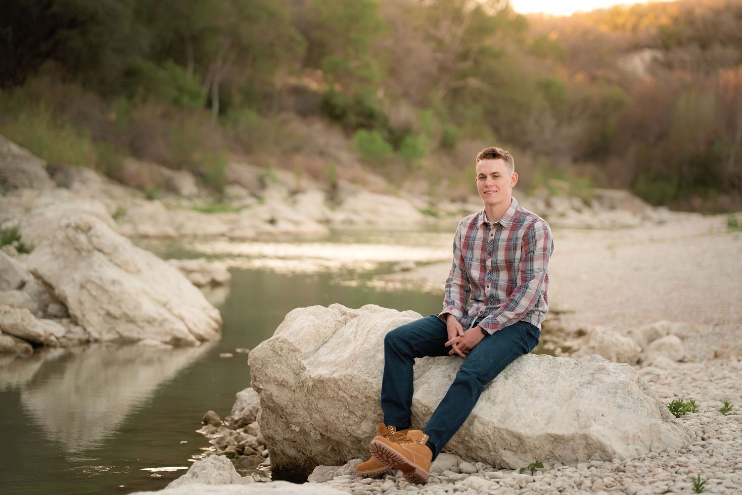 Young man in plaid shirt and jeans sitting on rocks by a serene river with lush green trees in background.