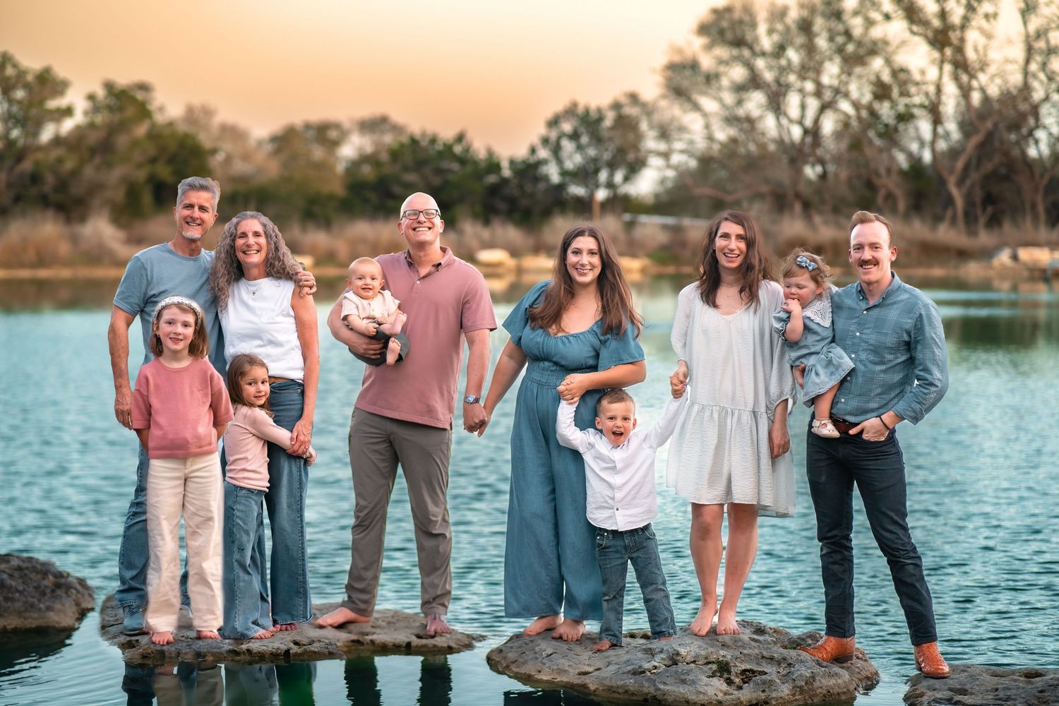 Multi-generational family portrait outdoors by a lake at golden hour, smiling adults and children on rocky shore.