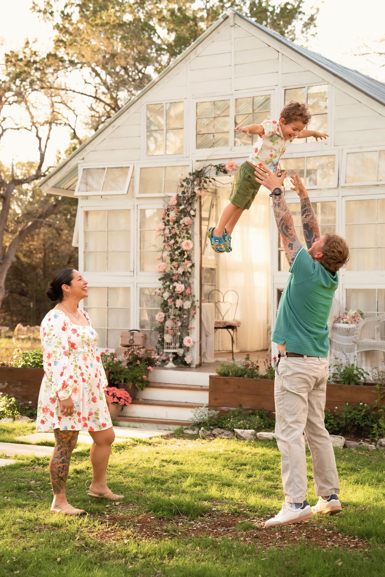 Family playing outdoors, father tossing child in air near white greenhouse with floral arch.
