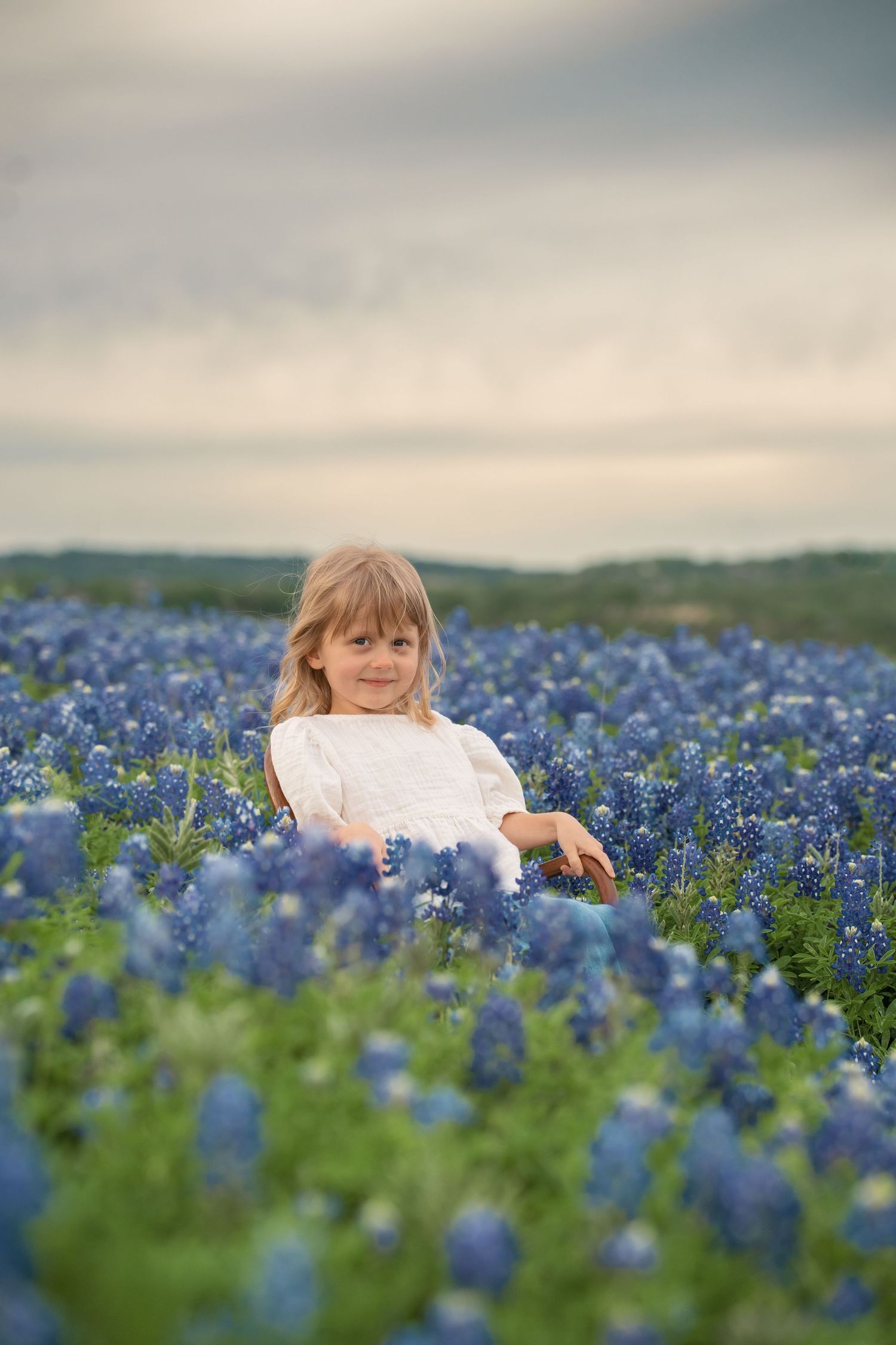 Young girl in white dress sitting in a field of blue Texas bluebonnet wildflowers under a cloudy sky.