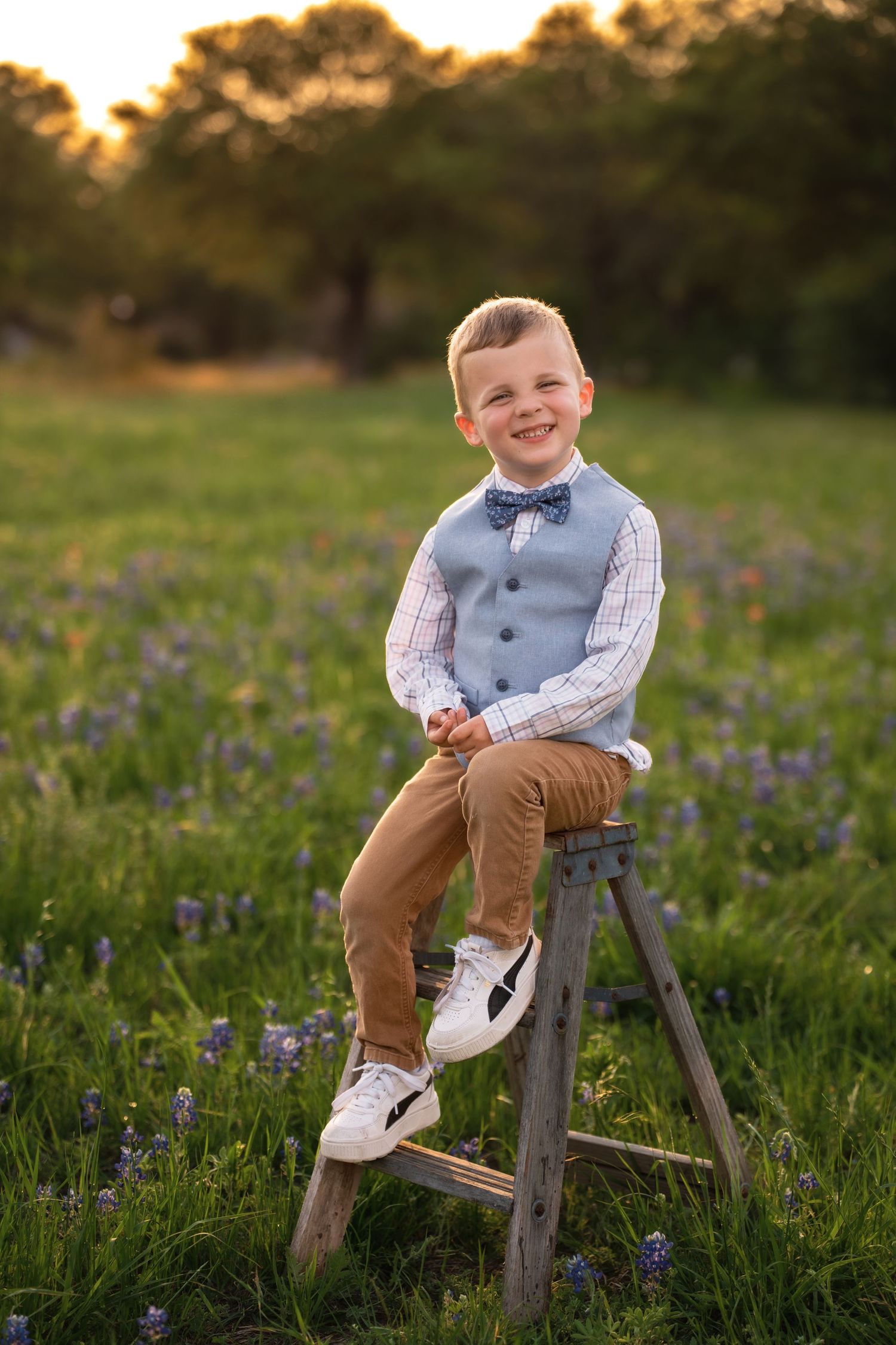 Child in gray vest and bow tie sits on wooden stool in wildflower field during golden hour sunset.