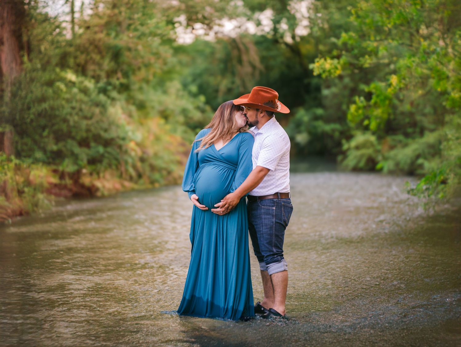 Pregnant couple sharing a tender kiss while standing in a shallow creek, woman in teal maternity gown, man in orange cowboy hat.