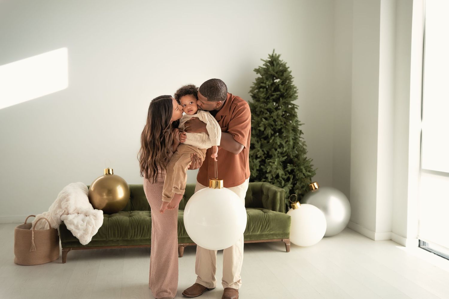 A family shares a tender kiss with their newborn near a decorated Christmas tree in a bright, modern living room.