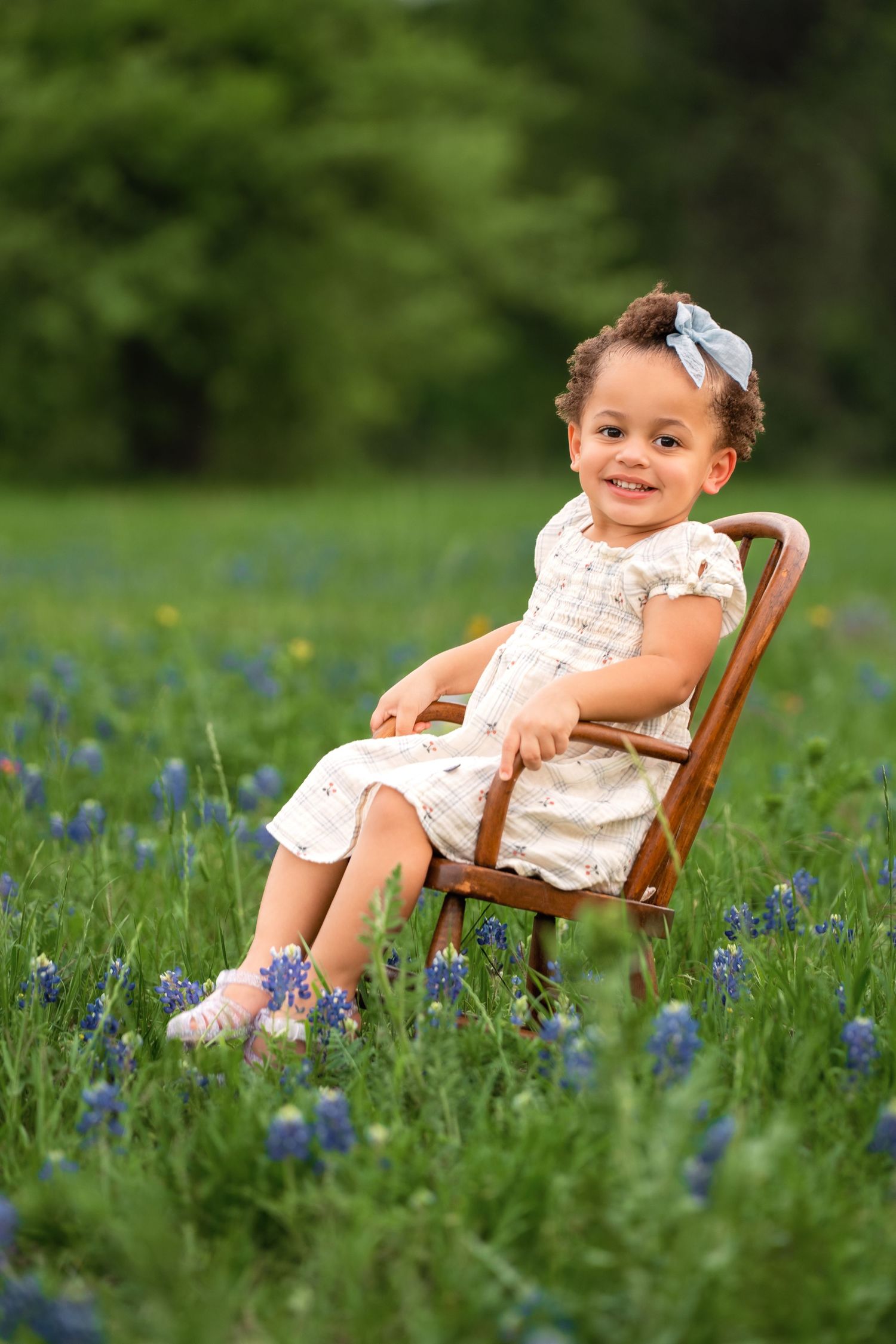 A small child sits in a wooden chair surrounded by blooming bluebonnets in a green spring meadow.