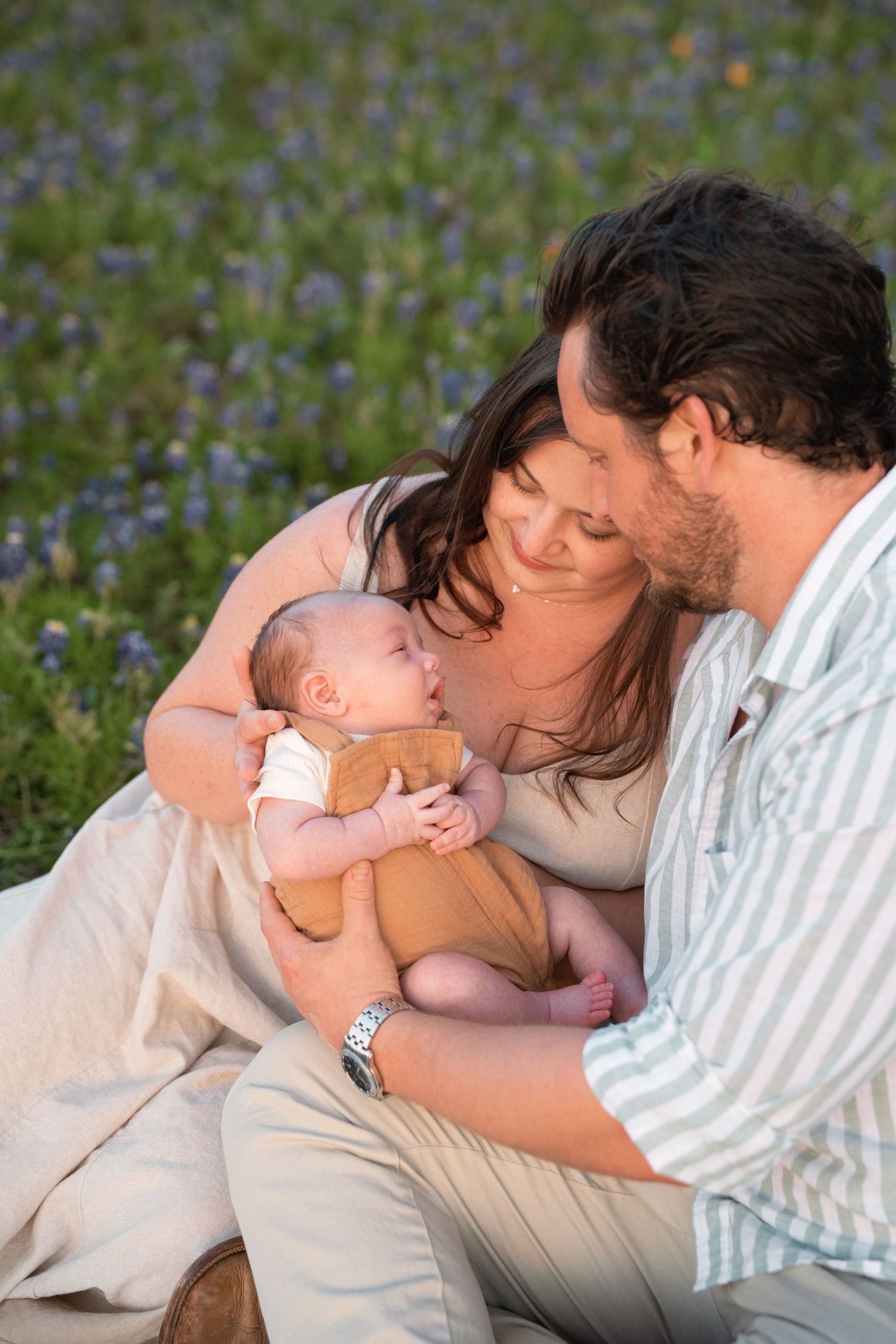 Parents share tender kisses with their baby while lying on a blanket among blooming bluebonnets in a spring meadow.
