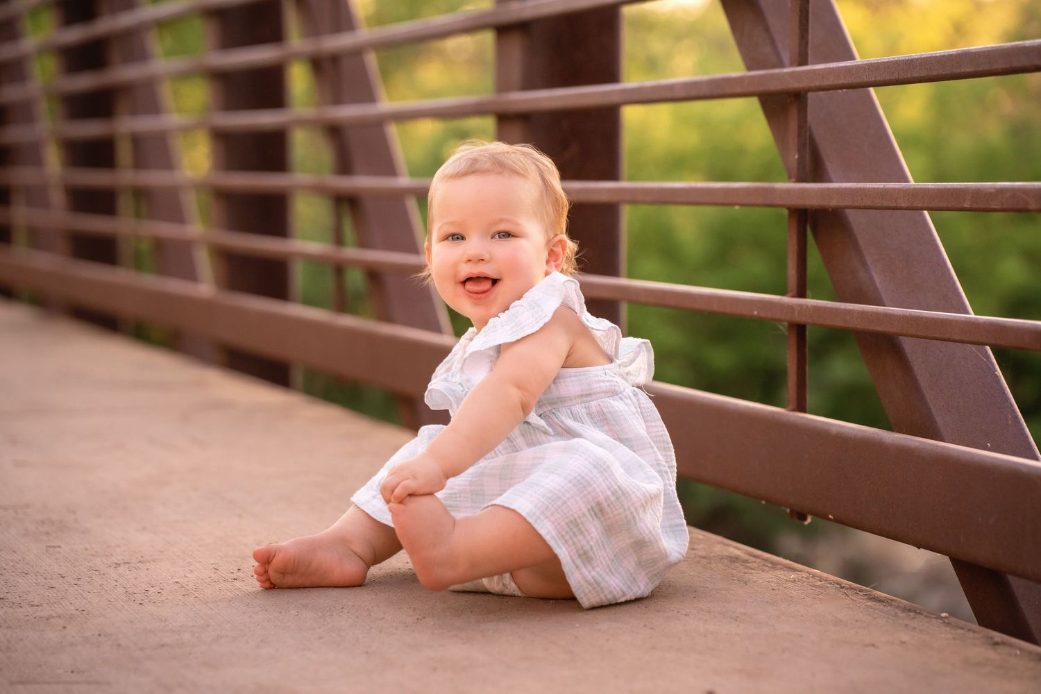Smiling baby girl in white dress sitting on a bridge outdoors in warm sunlight.