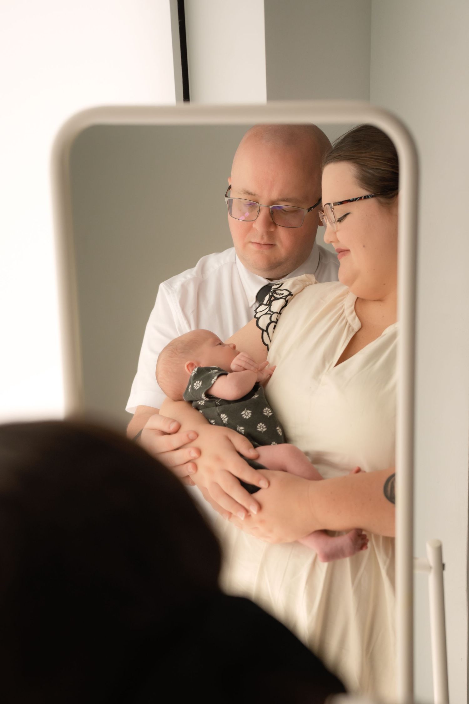 Couple with glasses admires their newborn baby together, captured in a mirror reflection.