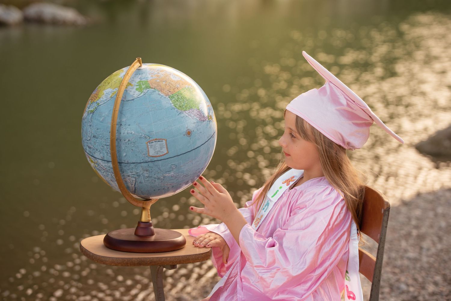 Young girl in pink graduation cap and gown examines a globe outdoors near a sunlit lake.