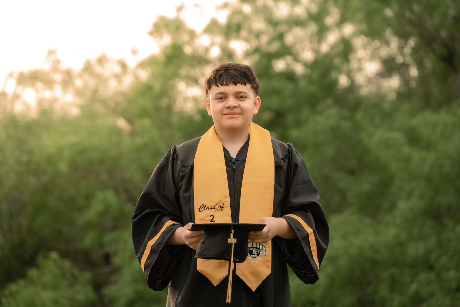 A young graduate in black cap and gown with gold stole poses outdoors among lush green trees at dusk.