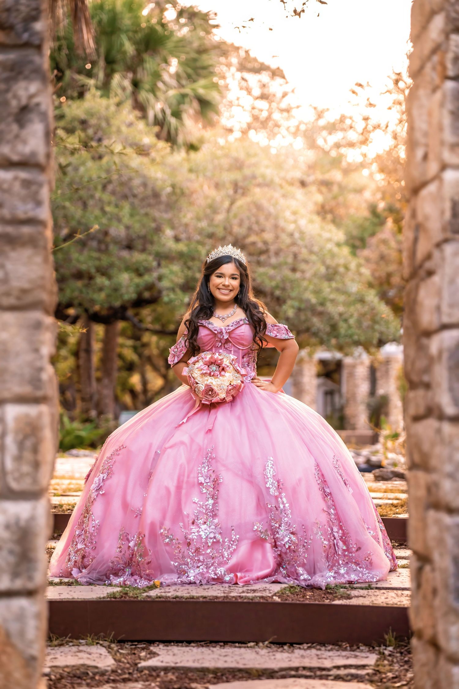 Young woman in pink quinceañera ball gown with floral embroidery and tiara holds bouquet outdoors.