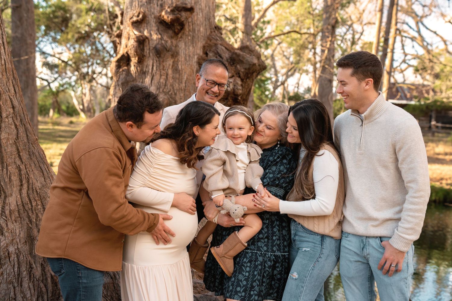 A joyful multigenerational family gathers outdoors near a large tree, smiling and admiring a happy baby in a warm, golden light.