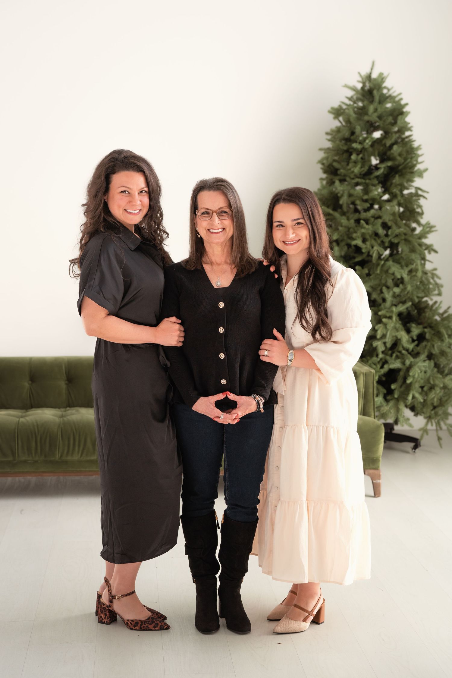 Three women pose together smiling in front of Christmas tree in bright white studio setting.