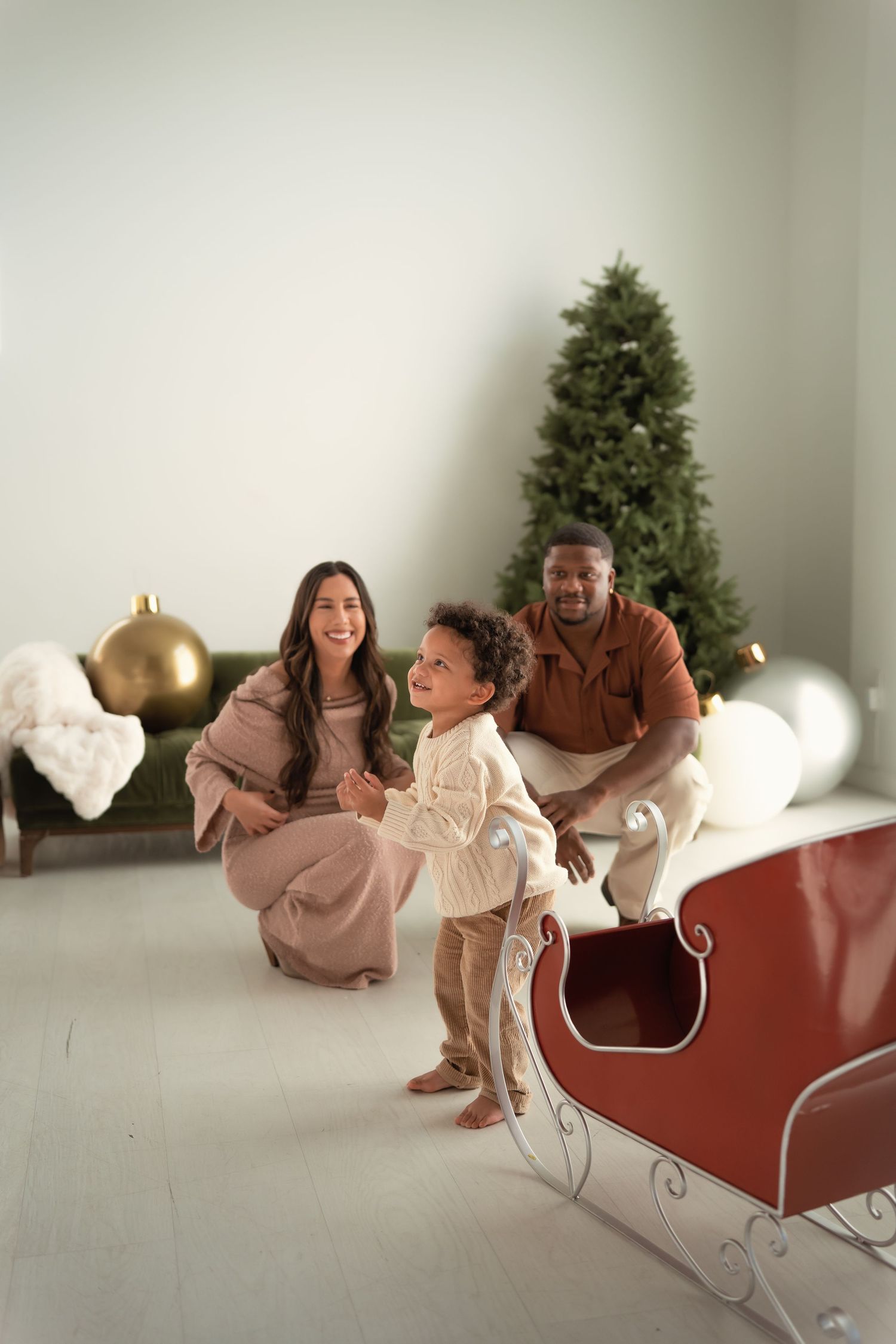 A joyful family with a toddler poses near a red sleigh and Christmas tree in a bright, festive holiday photo session.
