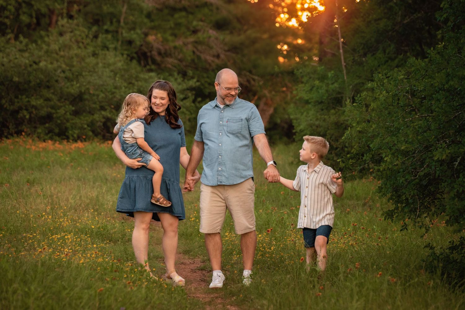 Family of four walking in a sunlit meadow at golden hour, parents holding hands with young children outdoors.
