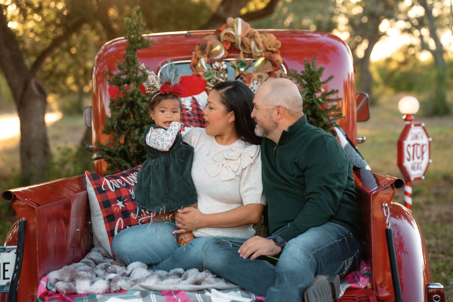 Family of three sitting in red truck bed with Christmas tree, celebrating holidays outdoors in festive attire.