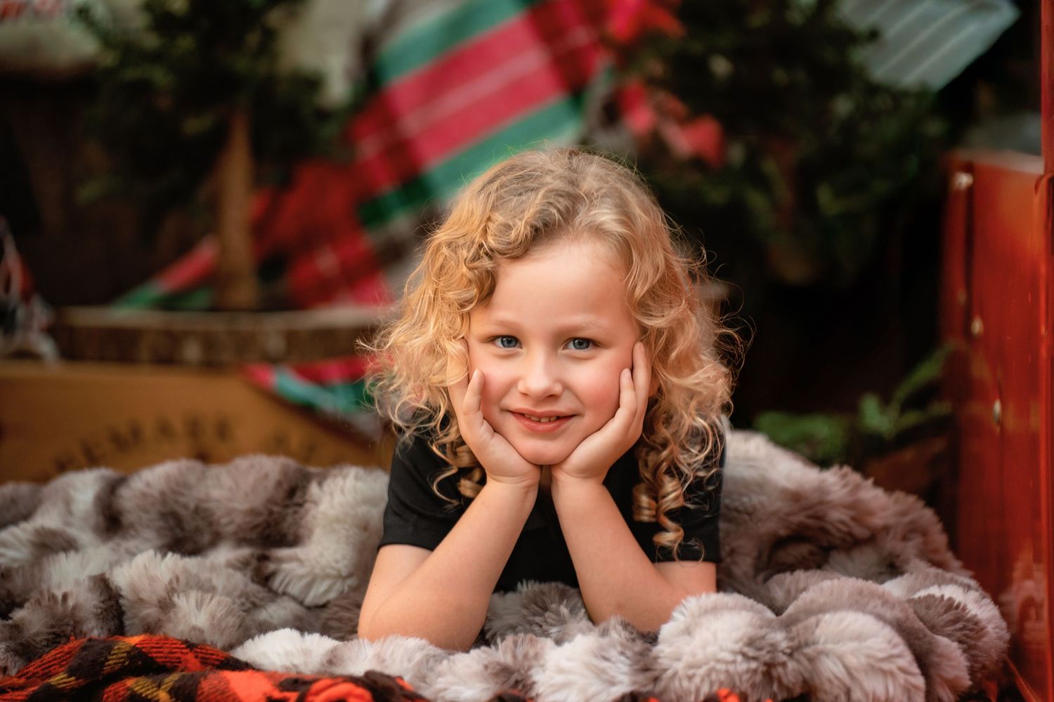 Smiling young girl with curly blonde hair resting chin on hands, surrounded by festive Christmas decorations.