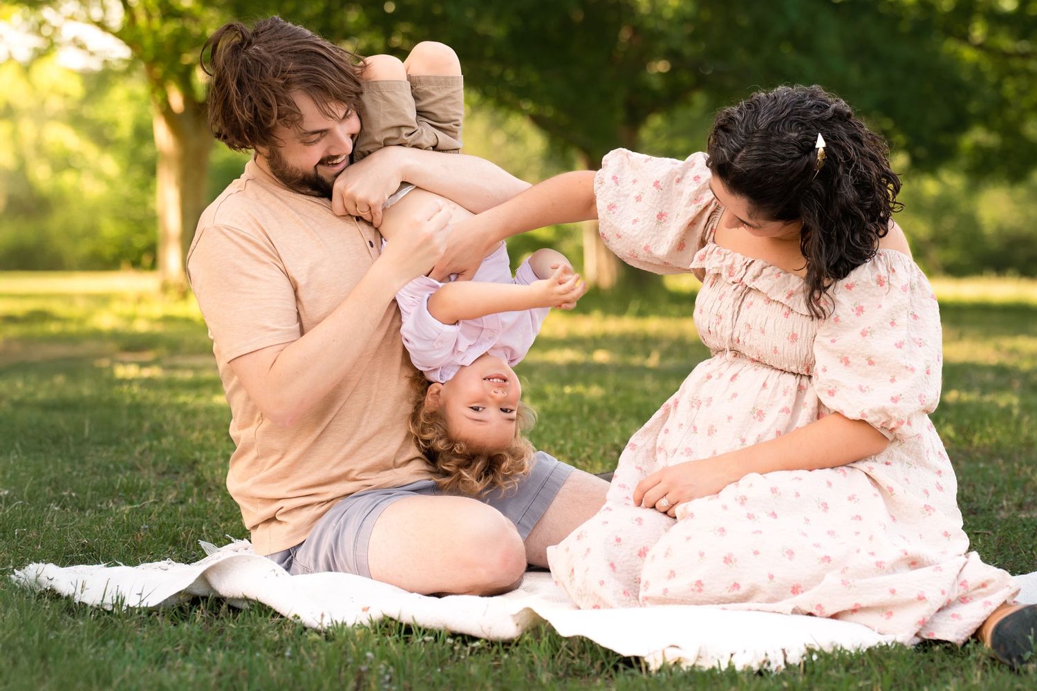 A happy family sitting on a blanket in a park, with a father playfully holding a baby upside down while the mother watches.