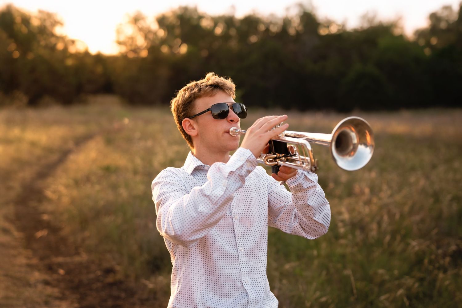 Young man in white shirt and sunglasses playing trumpet in a golden field at sunset.