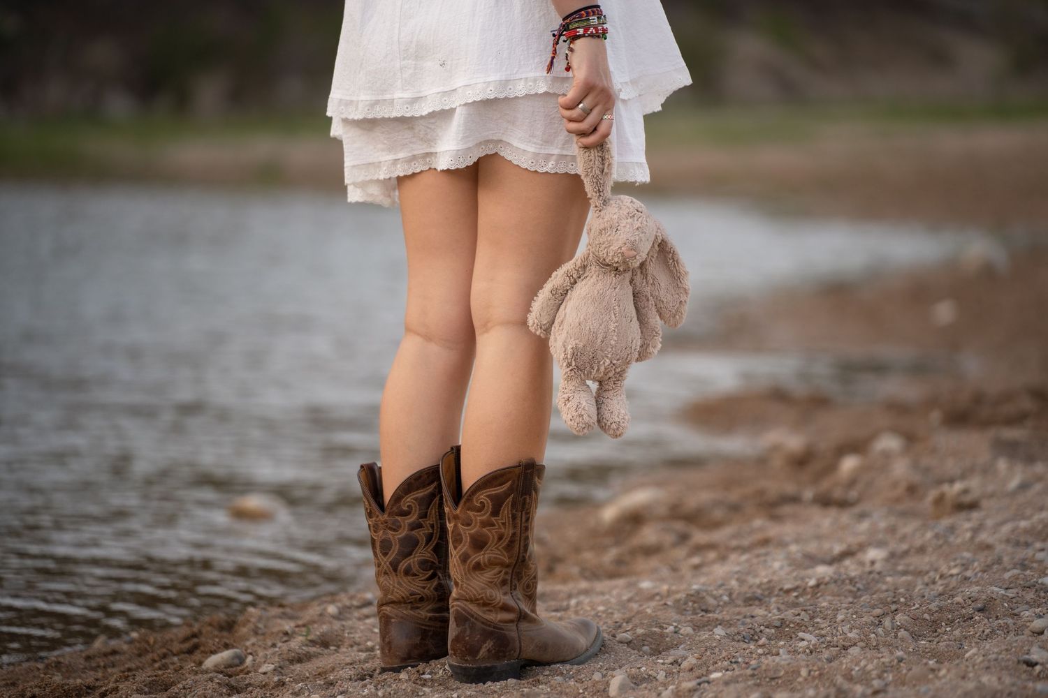 Woman in white dress and cowboy boots holds a stuffed bunny by a lakeside.