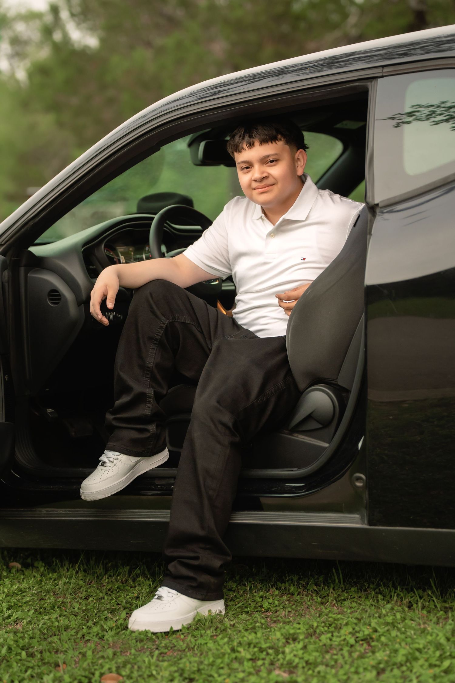 Young man in white polo and black pants sits relaxed in open car door of black vehicle outdoors.