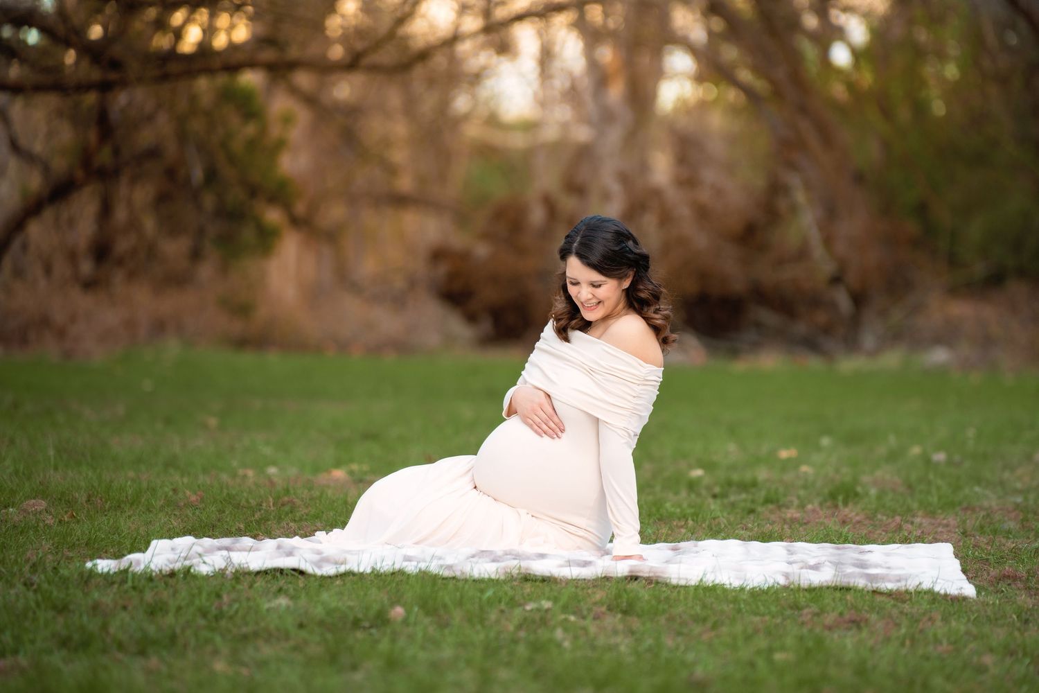 Pregnant woman in white off-shoulder gown sitting on grass in outdoor maternity photo session.