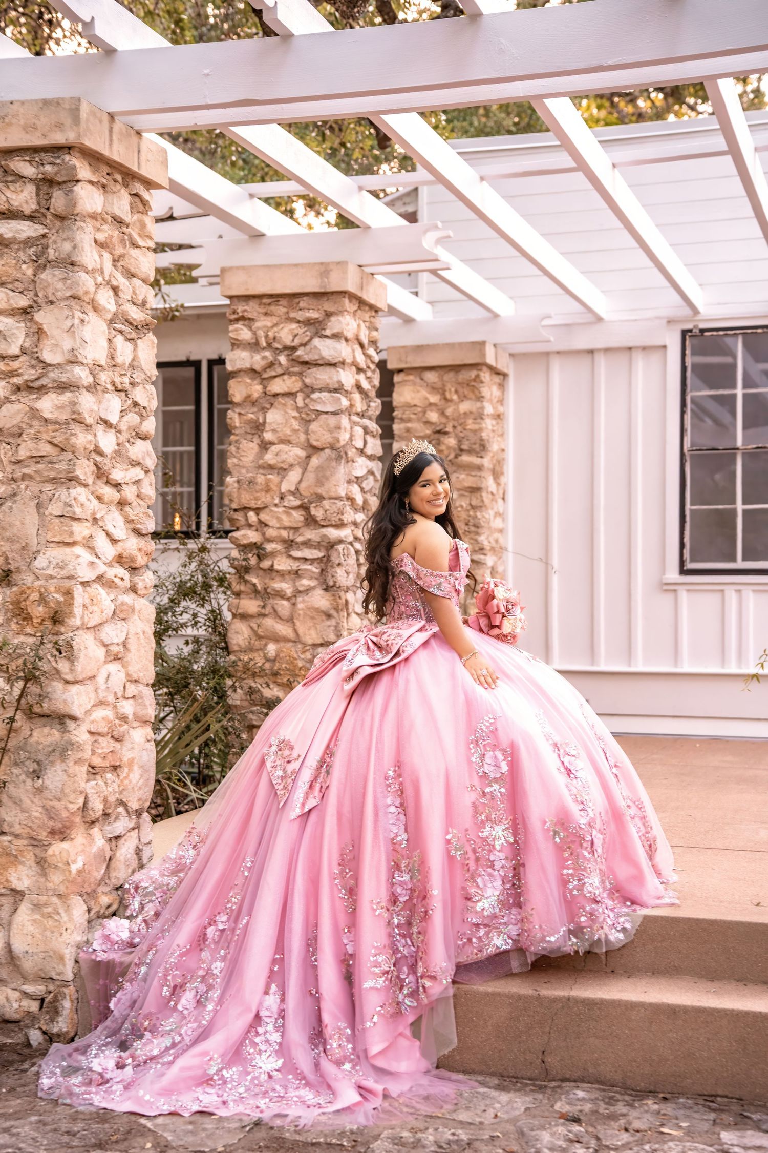 Elegant young woman in pink off-shoulder quinceañera gown poses on stone steps with bouquet.
