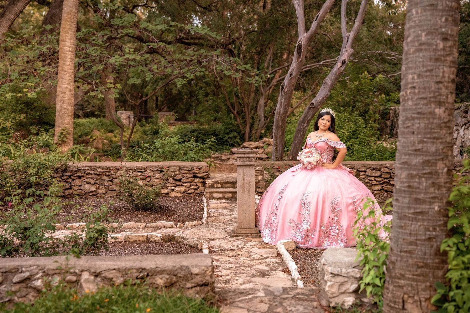 Young woman in pink quinceañera ball gown poses gracefully in a lush garden with stone walls and trees.
