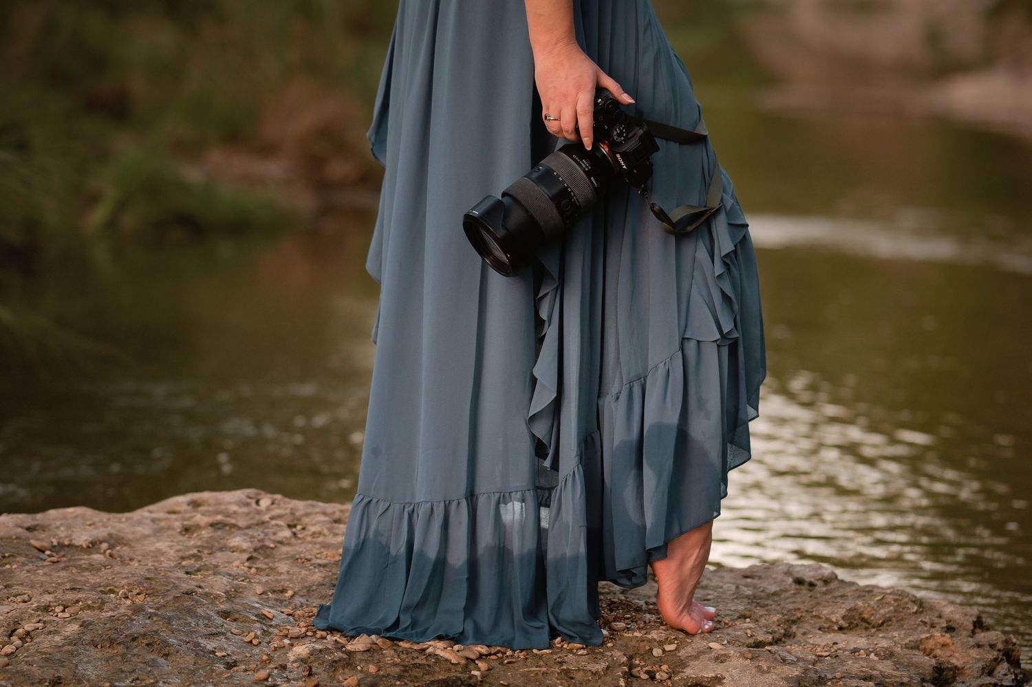 Woman in teal maxi dress holding DSLR camera standing barefoot on rocks near a river.