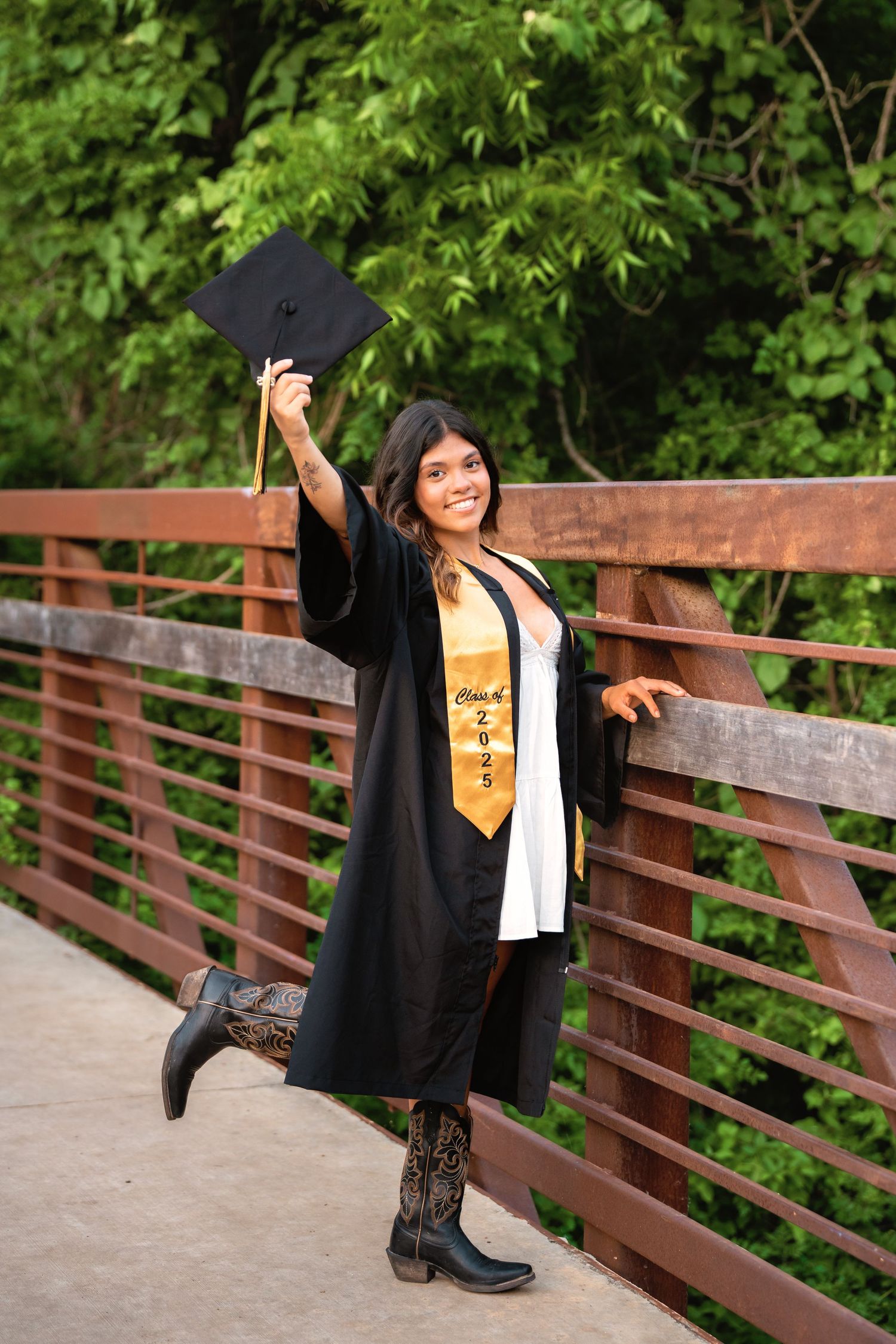 Smiling graduate in black gown and gold stole holds cap up on a bridge, wearing cowboy boots, class of 2024.