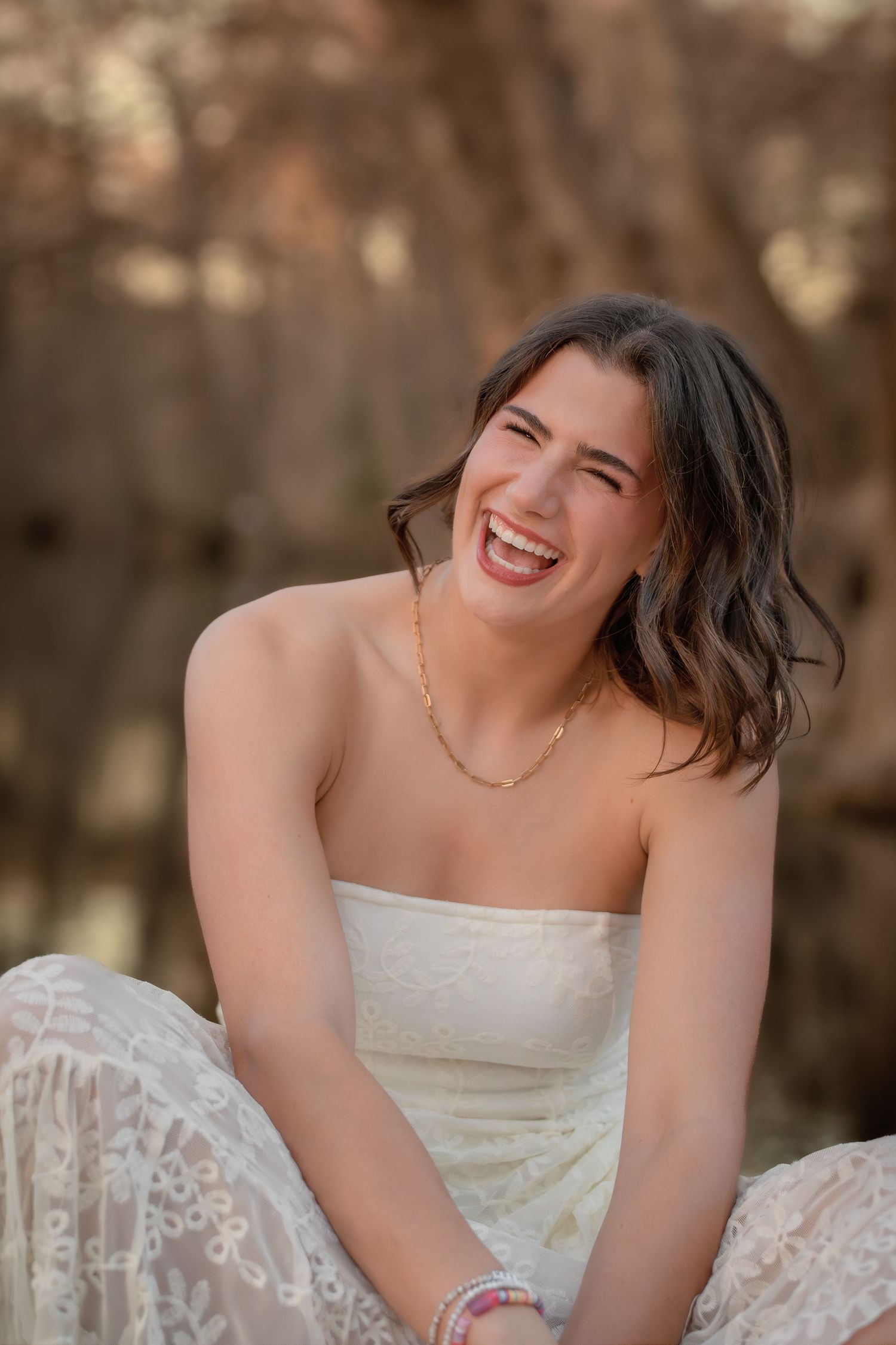 Laughing young woman in white strapless lace dress with gold necklace sitting outdoors.