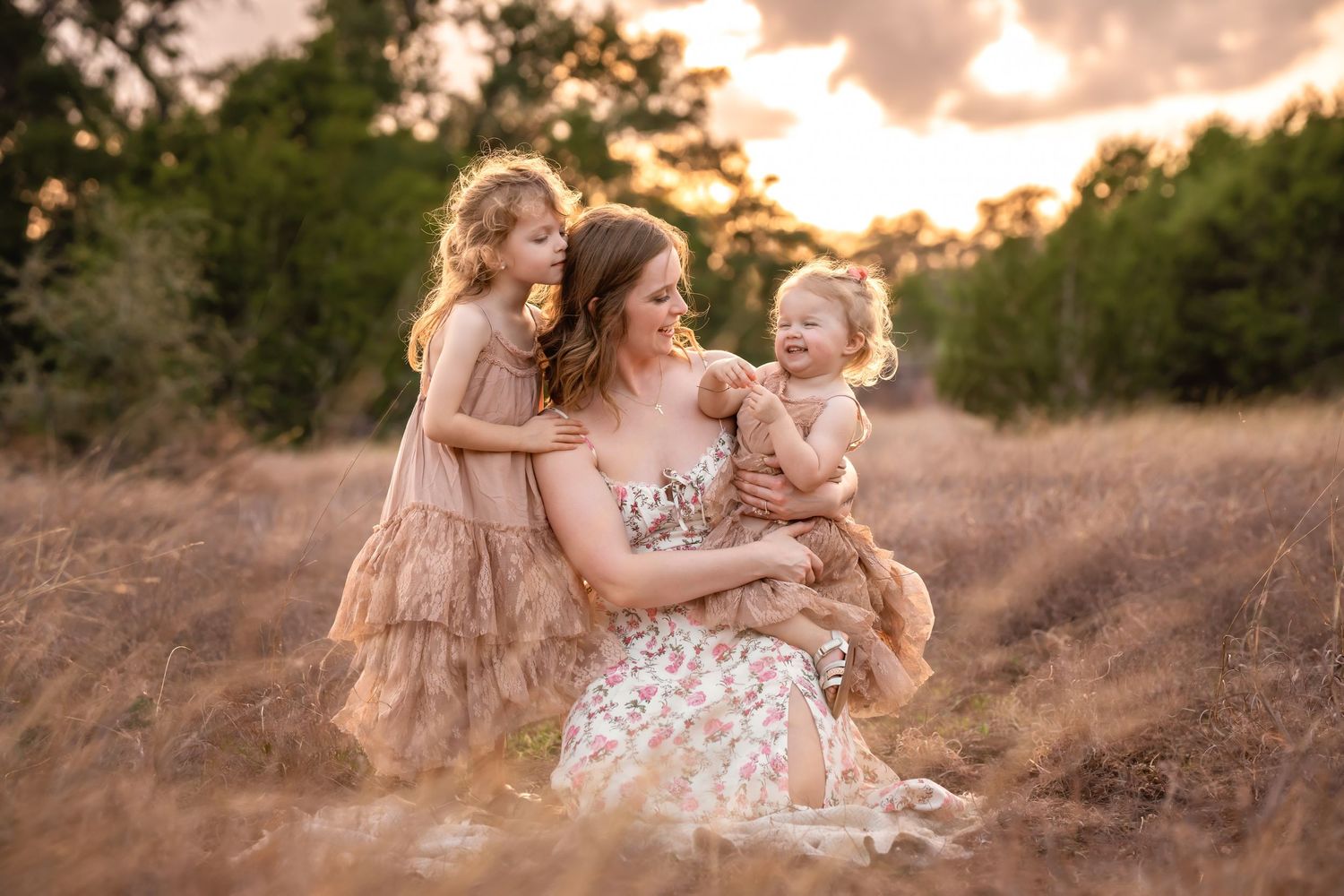 Mother sitting in golden field at sunset, smiling with two young daughters in floral and ruffled dresses.