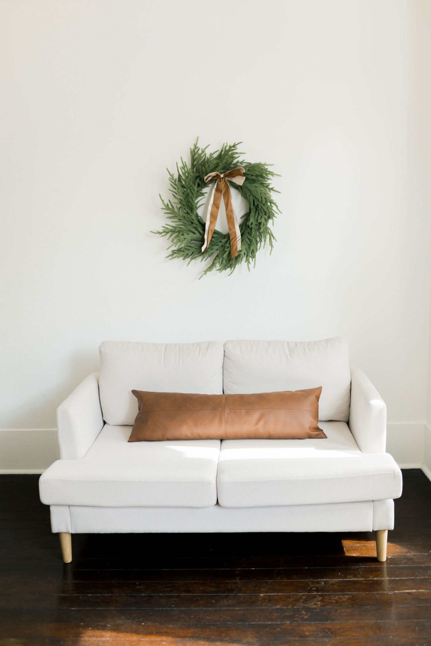 White loveseat sofa with brown leather pillow beneath a festive pine wreath with gold bow on white wall.