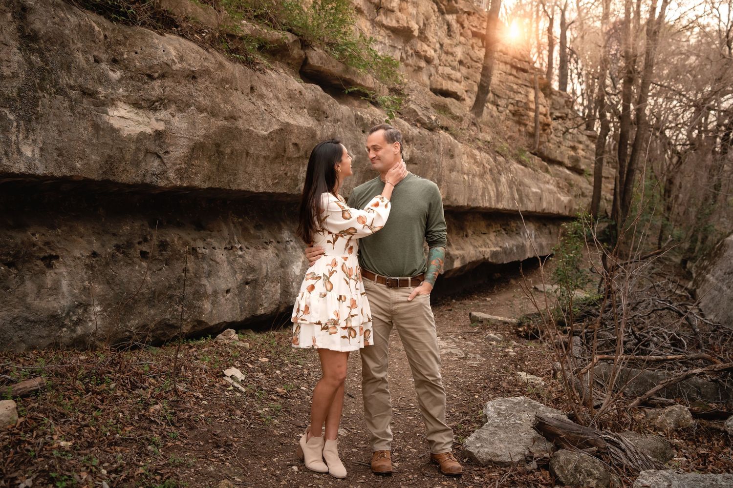 Couple embracing at golden hour near rocky cliffs in a forest, woman in floral dress, man in green sweater.