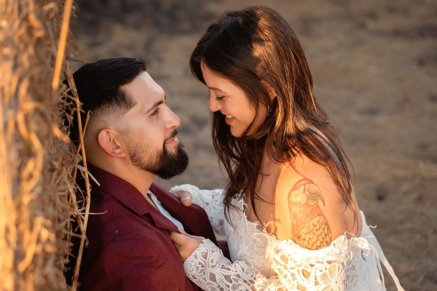 A couple shares a romantic moment leaning against a hay bale in golden sunset light, woman wearing lace dress with tattoo.