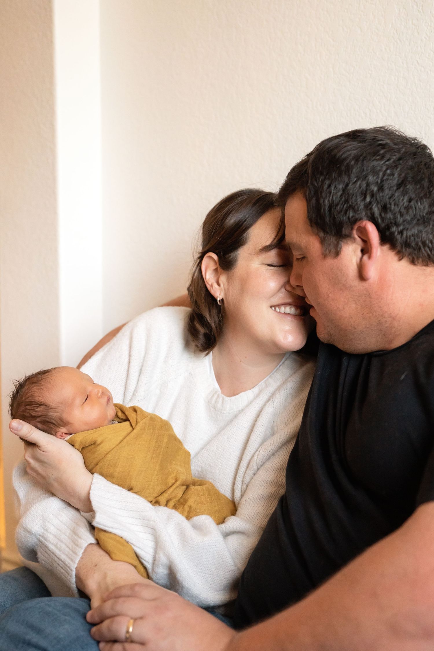 Happy couple sharing a tender moment while cradling their sleeping newborn baby wrapped in a mustard yellow swaddle.