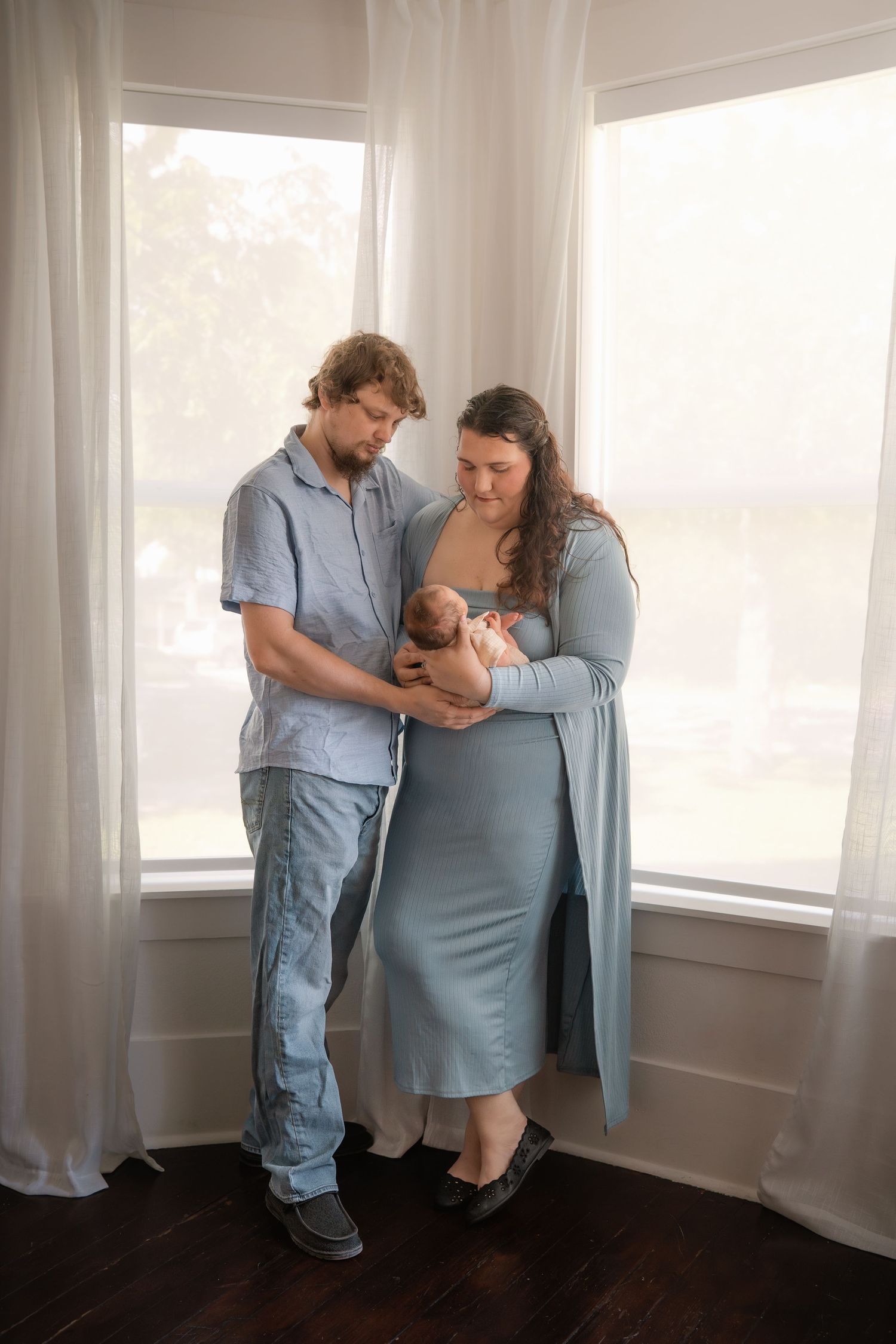 New parents stand by a bright window, lovingly gazing at their newborn baby, dressed in coordinating blue outfits.