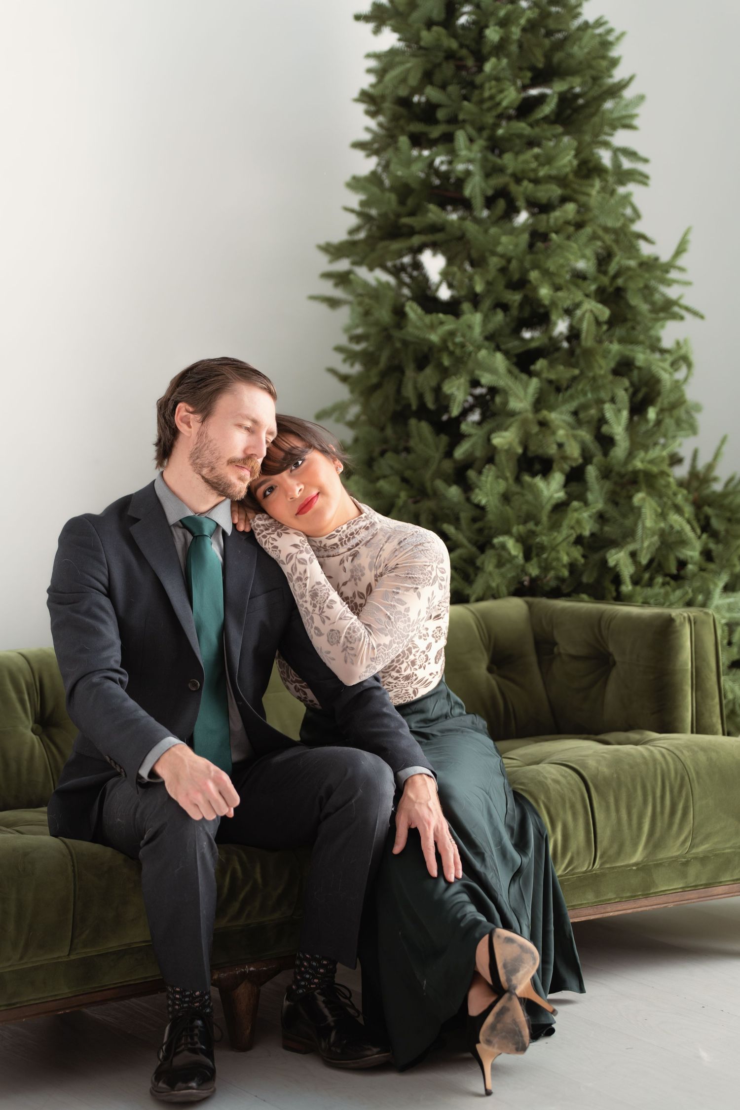 Couple sitting on green velvet sofa by Christmas tree, woman resting head on mans shoulder, dressed in elegant attire.