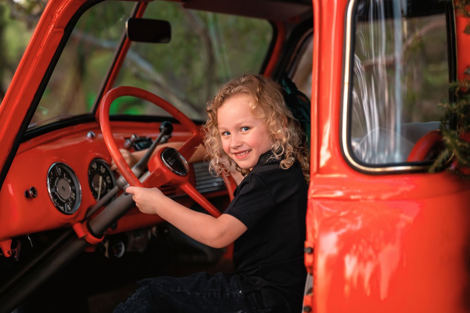 A smiling child with curly hair sits behind the steering wheel of a vintage red truck.