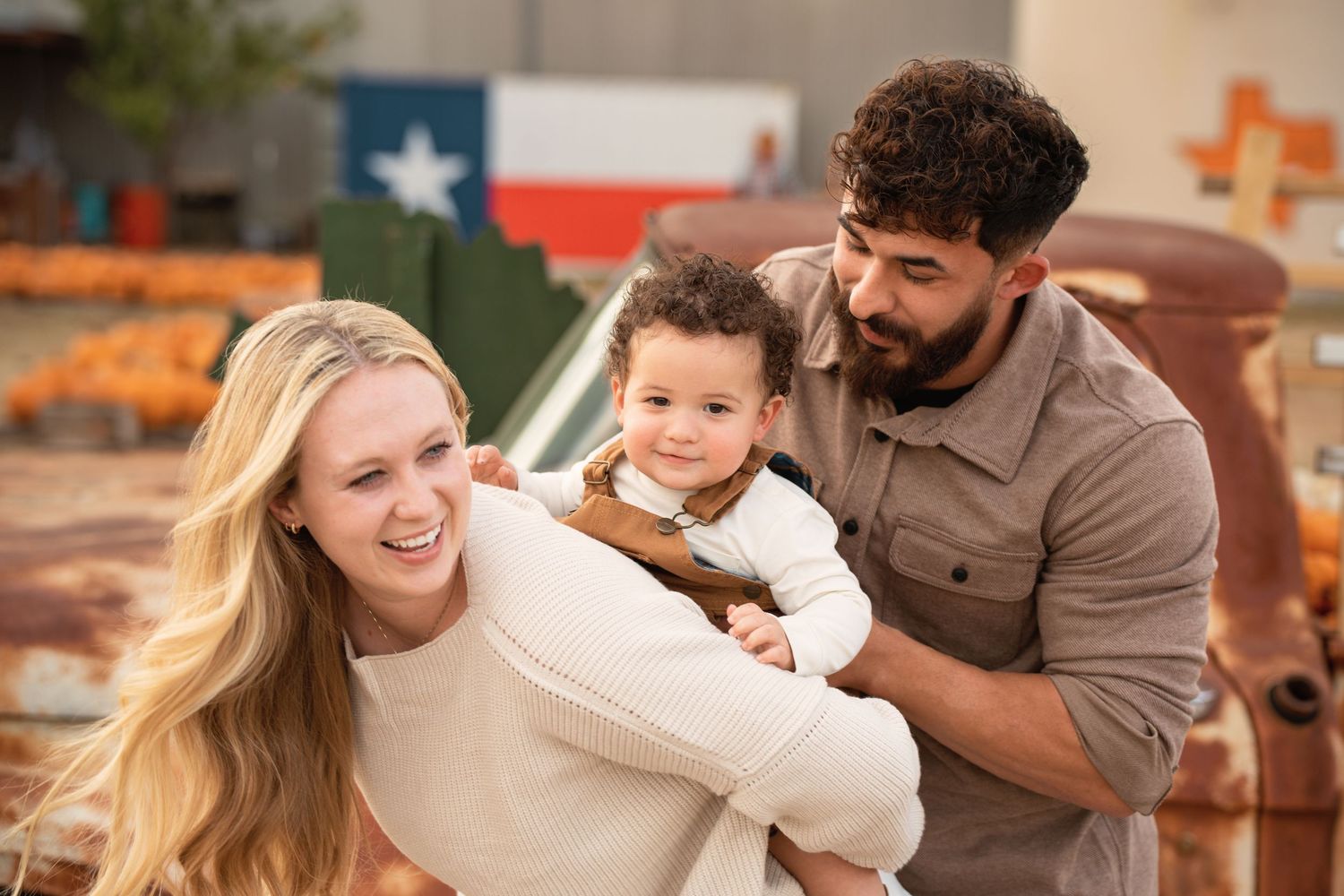 A happy family with a blonde mother, bearded father, and toddler posing near a Texas flag at a pumpkin patch.