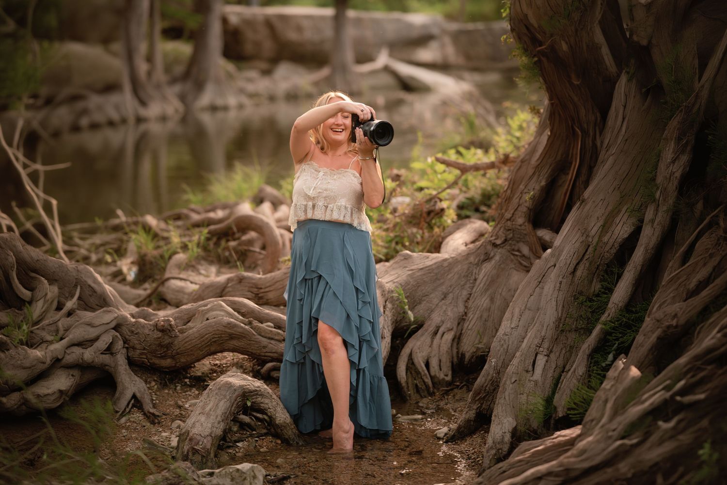 Woman in teal skirt photographing nature among twisted tree roots near water.